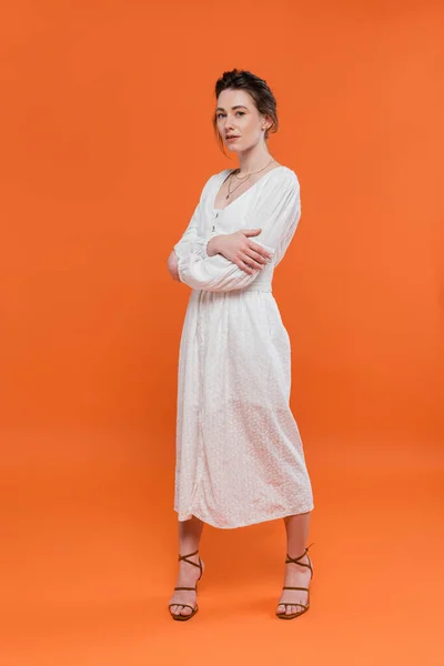 Tendências de verão, modelo em vestido de sol branco olhando para a câmera e de pé com os braços dobrados no fundo laranja, posando elegante, senhora em branco, fundo vibrante, moda, jovem — Fotografia de Stock