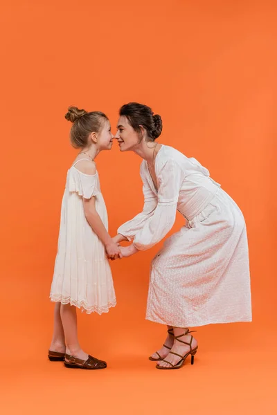 Summer trends, young mother holding hands with happy preteen daughter and standing on orange background, white sun dresses, togetherness, fashion and style concept, bonding and love, nose to nose — Stock Photo