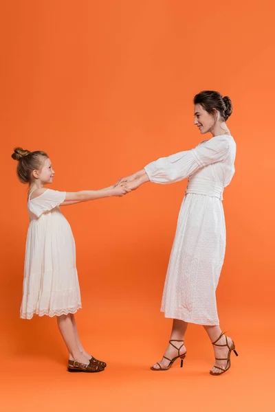 Summer trends, happy young mother holding hands with preteen daughter and standing on orange background, white sun dresses, togetherness, fashion and style concept, bonding and love — Stock Photo
