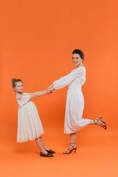 Summer trends, young mother holding hands with preteen daughter and standing on orange background, white sun dresses, togetherness, fashion and style concept, bonding, joyful — Stock Photo