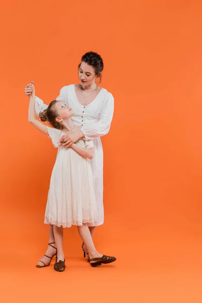 Sommertrends, Mutter hält Händchen mit frühgeborener Tochter und steht zusammen auf orangefarbenem Hintergrund, weiße Sonnenkleider, Zweisamkeit, Mode- und Stilkonzept, Bindung und Liebe — Stockfoto