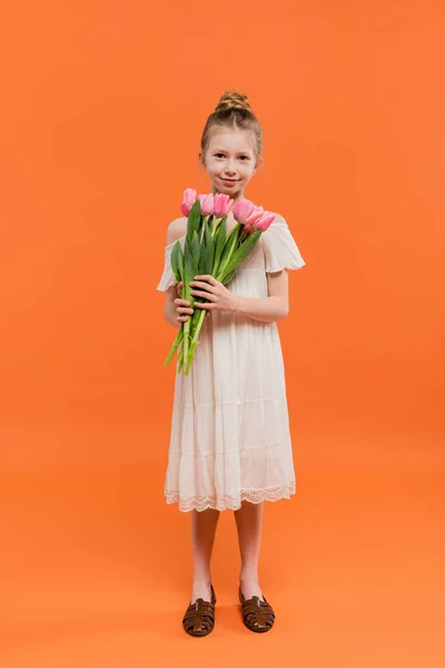Summer fashion, happy preteen girl in white sun dress holding pink tulips on orange background, fashion and style concept, bouquet of flowers, fashionable kid, vibrant colors, full length — Stock Photo