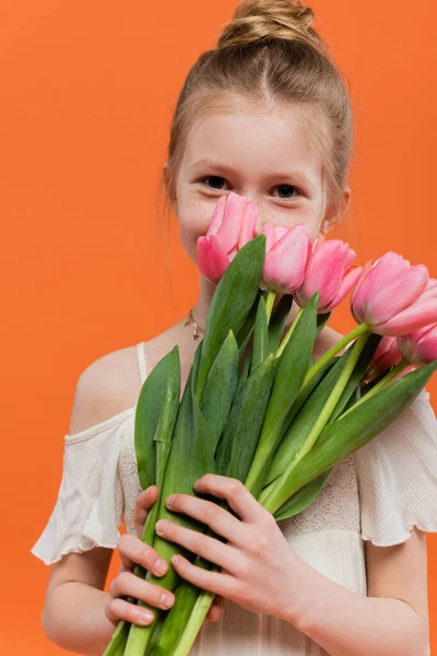 Niña preadolescente en vestido de sol blanco que sostiene tulipanes rosados sobre fondo naranja, concepto de moda y estilo, ramo de flores, niño de moda, colores vibrantes, cubriendo la cara - foto de stock