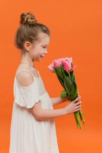 Side view of preteen girl in white sun dress holding pink tulips on orange background, fashion and style concept, bouquet of flowers, fashionable kid, vibrant colors, flowers and fashion — Stock Photo
