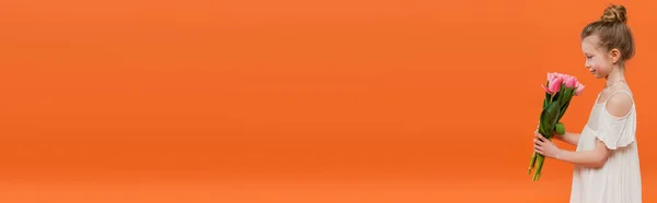 Vue latérale de la fille préadolescente en robe de soleil blanche tenant des tulipes roses sur fond orange, concept de mode et de style, bouquet de fleurs, enfant à la mode, couleurs vives, tenue d'été, bannière — Photo de stock