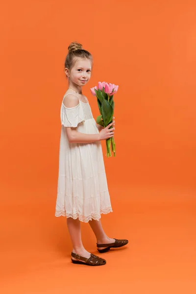 Summer dress, preteen girl in white sun dress holding pink tulips on orange background, fashion and style concept, bouquet of flowers, fashionable kid, vibrant colors, summer dress, full length — Stock Photo