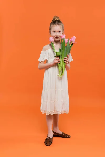 Colores vibrantes, niña preadolescente alegre en vestido de sol blanco que sostiene tulipanes rosados sobre fondo naranja, concepto de moda y estilo, ramo de flores, niño de moda, longitud completa - foto de stock