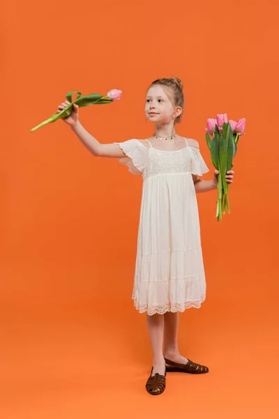 Blumenstrauß, frühgeborenes Mädchen in weißem Sonnenkleid mit rosa Tulpen auf orangefarbenem Hintergrund, Mode- und Stilkonzept, modisches Kind, lebendige Farben, Sommermode, niedliches Kind — Stockfoto