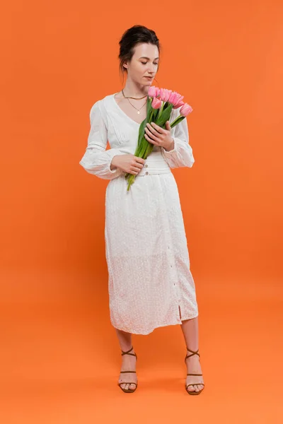 Buquê de flores, jovem mulher de vestido de sol branco segurando tulipas e de pé sobre fundo laranja, posando elegante, senhora de branco, fundo vibrante, moda, verão, comprimento total — Fotografia de Stock