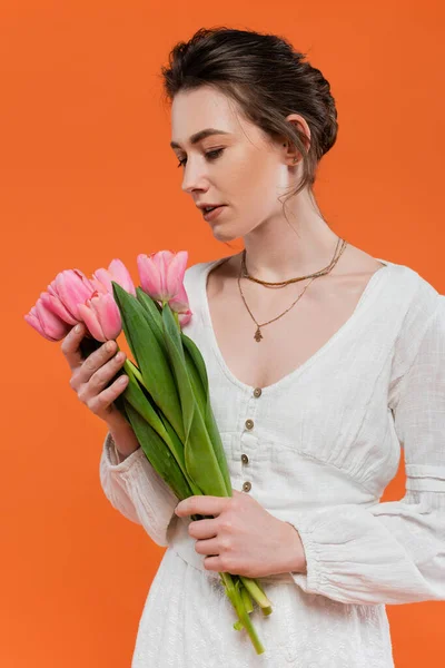 Ramo de flores, mujer joven en vestido de sol blanco con tulipanes y de pie sobre fondo naranja, posando con estilo, dama de blanco, fondo vibrante, moda, verano - foto de stock