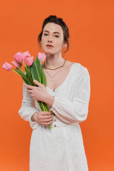 Buquê de flores, atraente jovem mulher em vestido de verão branco segurando tulipas e de pé sobre fundo laranja, posando elegante, senhora em branco, fundo vibrante, moda, verão — Fotografia de Stock