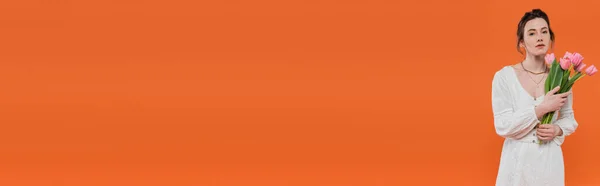 Buquê de flores, jovem mulher em vestido de sol segurando tulipas e de pé sobre fundo laranja, posando elegante, senhora em branco, fundo vibrante, moda, verão, banner — Fotografia de Stock