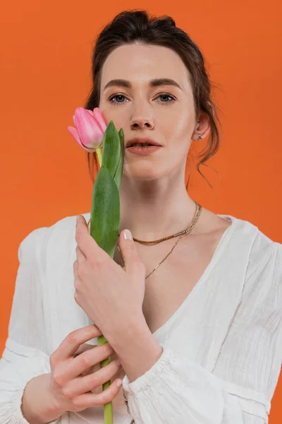 Moda cotidiana, mujer joven en vestido de sol blanco sosteniendo tulipán rosa y de pie sobre fondo naranja, dama en blanco, fondo vibrante, moda y naturaleza, verano, retrato - foto de stock