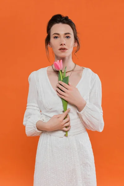 Sensualidad, moda cotidiana, mujer joven en vestido de sol blanco con tulipán rosa y de pie sobre fondo naranja, dama de blanco, fondo vibrante, moda y naturaleza, verano, retrato - foto de stock