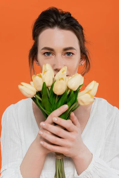 Bouquet de fleurs, mode quotidienne, jeune femme en robe de soleil blanche tenant des tulipes jaunes et debout sur fond orange, dame en blanc, fond vibrant, mode, été — Photo de stock