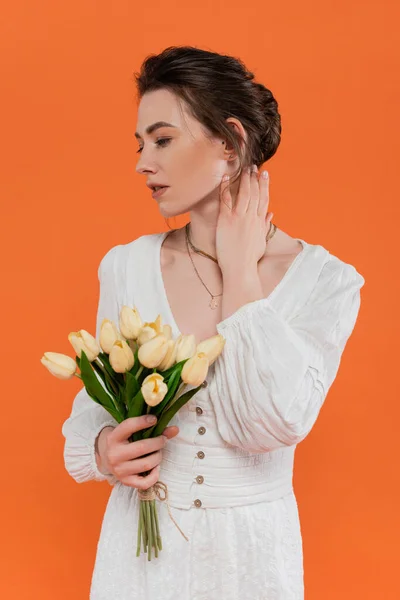 Concepto de moda, mujer joven en vestido de sol blanco con tulipanes amarillos y de pie sobre fondo naranja, dama en blanco, fondo vibrante, moda, verano, ramo de flores - foto de stock
