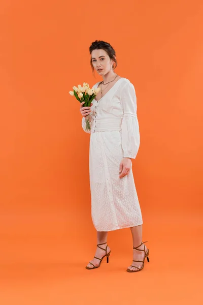 Concepto de moda, mujer bastante joven en vestido de sol blanco sosteniendo tulipanes amarillos y de pie sobre fondo naranja, dama en blanco, fondo vibrante, moda, verano, ramo de flores, longitud completa - foto de stock
