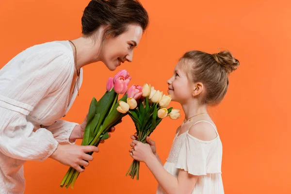 Счастливая мать и ребенок с цветами, молодая женщина и девочка, держащие тюльпаны и глядя друг на друга на оранжевом фоне, летняя мода, солнечные платья, женские связи — стоковое фото