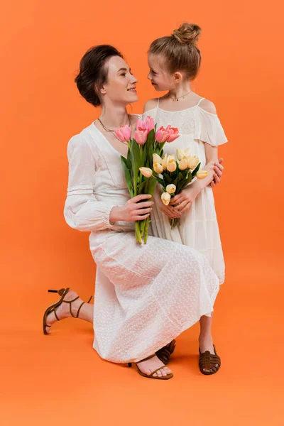Счастливая мать и дочь с цветами, молодая женщина и девочка, держа тюльпаны и позируя на оранжевом фоне, летняя мода, солнечные платья, женские связи — стоковое фото