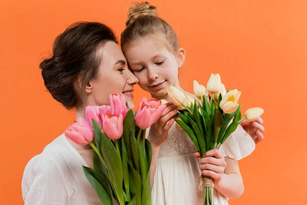 Happy mother and daughter with tulips, young woman and girl holding flowers and posing on orange background, summer fashion, sun dresses, female bonding, family love — Stock Photo