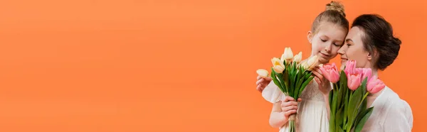 Alegre madre e hija con tulipanes, joven mujer y niña sosteniendo flores y posando sobre fondo naranja, moda de verano, vestidos de sol, vinculación femenina, amor familiar, bandera - foto de stock