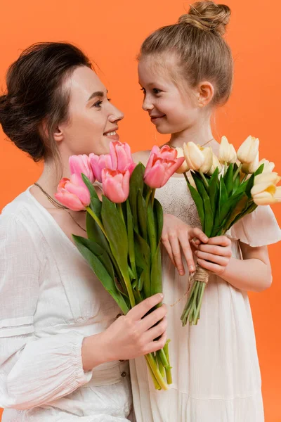 Happy mother and daughter with tulips, young woman and girl holding flowers and looking at each other on orange background, summer fashion, stylish family, sensuality — Stock Photo