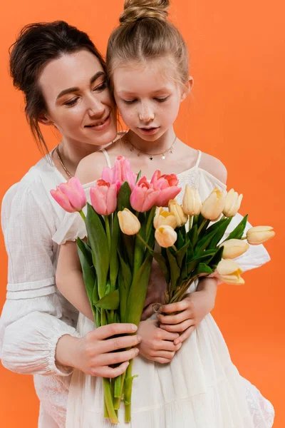 Happy mother and daughter with tulips, young woman and girl holding flowers and posing on orange background, summer fashion,  family love, sun dresses, female bonding — Stock Photo