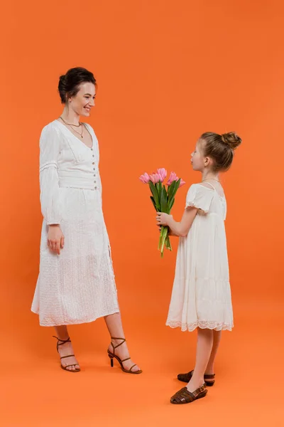 Mother`s day, cute preteen girl holding bouquet of flowers near mother on orange background, bonding, white dresses, pink tulips, happy holiday, vibrant colors, joyful occasion — Stock Photo