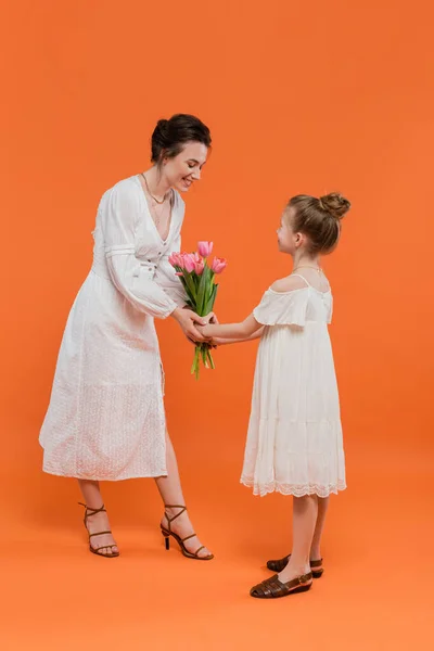 Muttertag, süßes Frühchen, das der Mutter einen Blumenstrauß auf orangefarbenem Hintergrund schenkt, Bindung, weiße Kleider, rosa Tulpen, frohe Feiertage, lebendige Farben, freudige Gelegenheit — Stockfoto