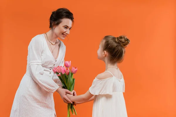 Mother`s day, preteen girl giving bouquet of flowers to smiling mother on orange background, bonding, white dresses, pink tulips, happy holiday, vibrant colors, joyful occasion — Stock Photo