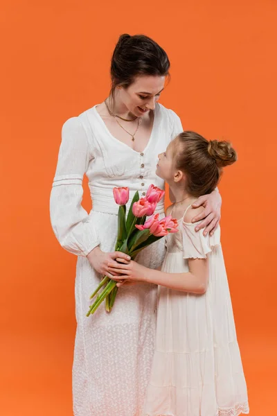 Mother`s day, mother hugging preteen daughter with bouquet of flowers on orange background, bonding, white dresses, pink tulips, happy holiday, vibrant colors, joyful occasion — Stock Photo