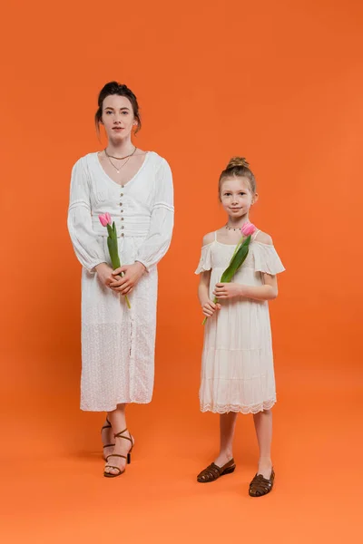 Heureuse mère et fille avec des tulipes, jeune femme et fille tenant des fleurs et debout sur fond orange, style familial, occasion joyeuse, mode et nature — Photo de stock