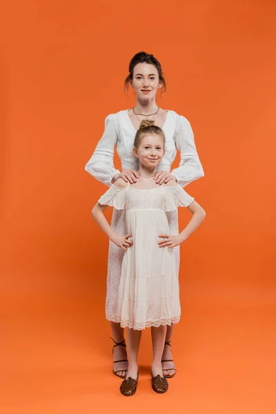 Modern parenting, summer fashion, woman hugging preteen daughter and standing together in white sun dresses on orange background, female bonding, fashionable family, summer style — Stock Photo