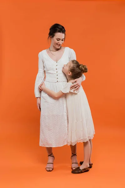 Weibliche Bindung, fröhliche Frühchen Mädchen umarmt Mutter auf orangefarbenem Hintergrund, volle Länge, Glück, weiße Sonnenkleider, Sommermode, Zweisamkeit, Liebe, Frauen-Stil, moderne Elternschaft — Stockfoto