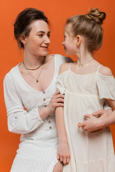 Motherly love, pretty mother smiling and looking at daughter on orange background, white sun dresses, summer fashion, togetherness, love, female bonding, women style, modern parenting — Stock Photo