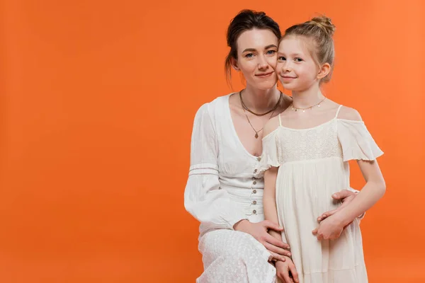 Motherly love, happy young woman hugging preteen girl on orange background, white sun dresses, summer fashion, togetherness, love, female bonding, mother and daughter, modern parenting — Stock Photo