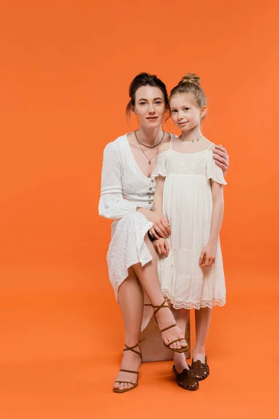 Motherly love, brunette young woman sitting on chair and hugging preteen daughter on orange background, white sun dresses, summer fashion, togetherness, love, female bonding, modern parenting — Stock Photo