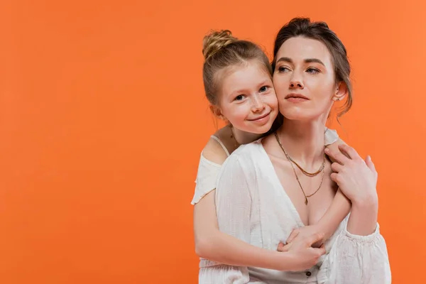 Мати і дочка, весела дівчина, що обіймає молоду жінку на помаранчевому фоні, білі сонцезахисні сукні, сучасне батьківство, літня мода, непохитність, любов — стокове фото