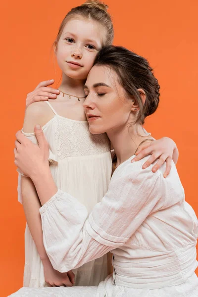 Motherly love, young woman embracing her daughter on orange background, closed eyes, modern parenting, family fashion, white sun dresses, togetherness, love, female bonding — Stock Photo