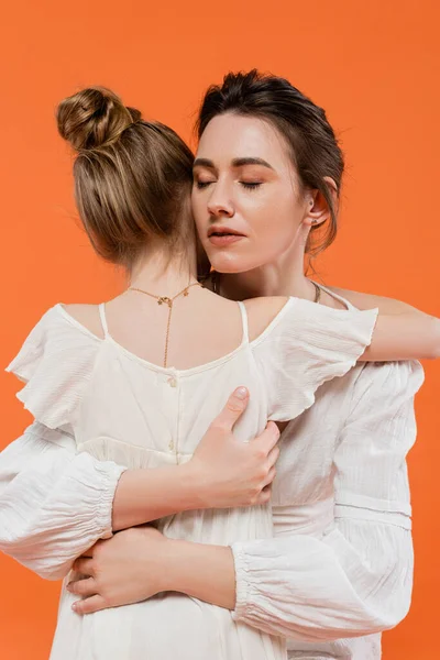 Motherly love, caring mother hugging daughter on orange background, closed eyes, white sun dresses, female bonding, modern parenting, love and care, support, togetherness — Stock Photo