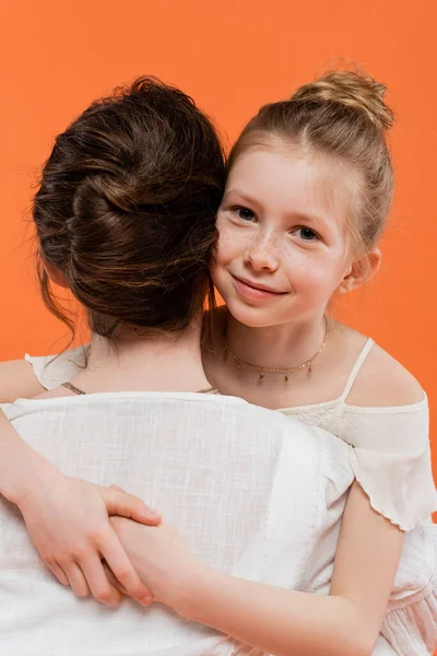 Семейные моменты, счастливая девочка-подросток обнимает брюнетку мать на оранжевом фоне, глядя на камеру, белые солнечные платья, женские связи, любовь и забота, единение, радость — стоковое фото