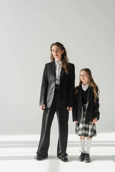 Работающая мать и школьница, веселая девочка в школьной форме, стоящая с деловой женщиной в костюме на сером фоне, держась за руки, официальный наряд, модная семья, сближение, современное воспитание — стоковое фото