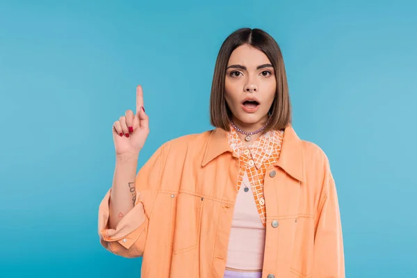 Summer fashion, shocked young woman with short hair and piercing in nose and tattoos posing in casual outfit on blue background, everyday makeup, orange shirt, generation z, showing and pointing up — Stock Photo