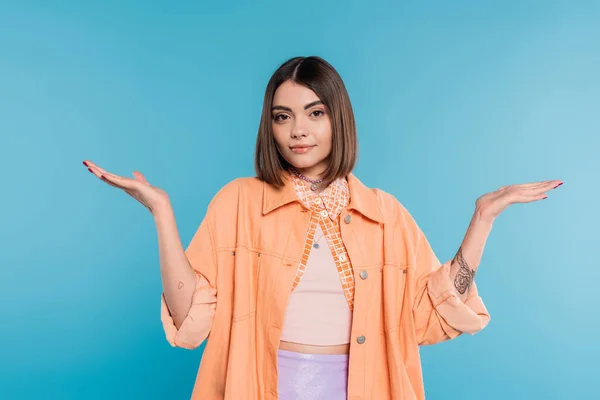 Summer outfit, brunette young woman with short hair and piercing in nose and tattoos posing in casual outfit on blue background, orange shirt, generation z, not knowing and gesturing with hands — Stock Photo