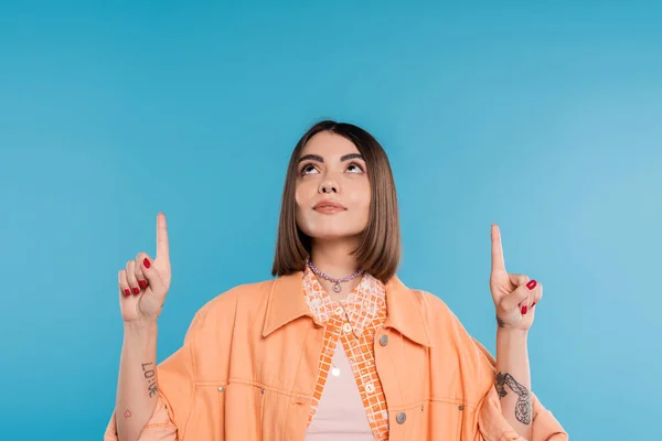 Summer fashion, brunette young woman with short hair and piercing in nose and tattoos posing in casual outfit on blue background, everyday makeup, orange shirt, generation z, showing and pointing up — Stock Photo
