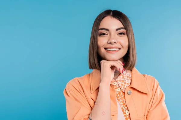 Summer fashion, cheerful young woman with short hair and piercing in nose and tattoos looking at camera on blue background, everyday makeup, orange shirt, generation z — Stock Photo