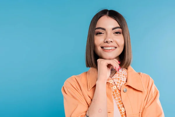 Mode d'été, jeune femme gaie aux cheveux courts et percing dans le nez et tatouages regardant la caméra sur fond bleu, maquillage quotidien, chemise orange, génération z — Photo de stock