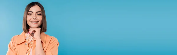 Mujer alegre, modelo de moda morena con pelo corto, piercing en la nariz y tatuajes posando en traje casual sobre fondo azul, maquillaje diario, camisa naranja, generación z, pancarta - foto de stock
