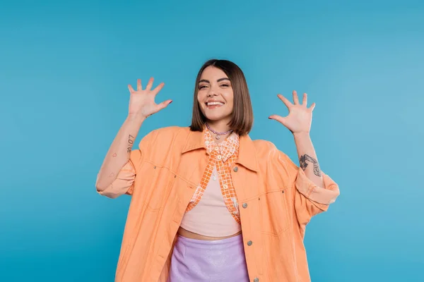 Excited young woman gesturing and smiling while looking at camera, on blue background, summer outfit, generation z, short brunette hair, orange shirt, pierced nose, tattooed — Stock Photo