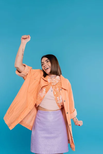 Summer outfit, excited young woman smiling while gesturing on blue background, summer outfit, generation z, short brunette hair, orange shirt, pierced nose, tattooed — Stock Photo